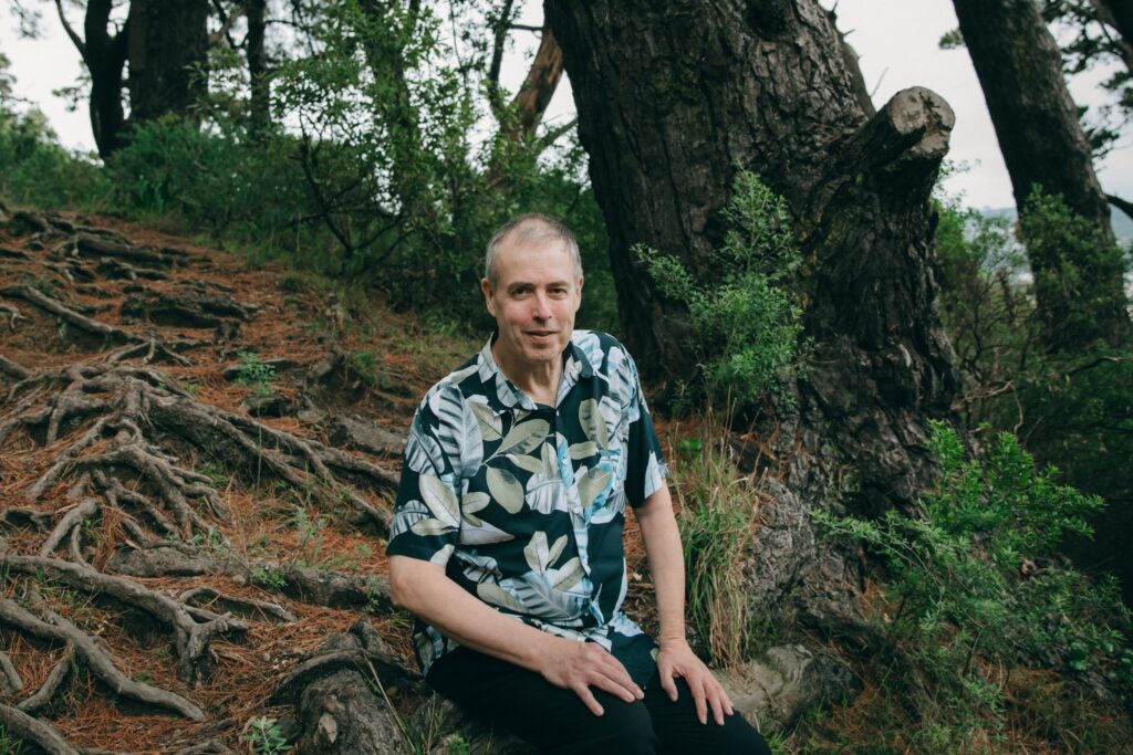 Photo of Tim Jones sitting on a tree root in a forest. Behind him are undergrowth, pine trees, and grass, with a distant skyline in the background. Upslope are roots, pine needles, and grass. The author is wearing a flower-patterned shirt.